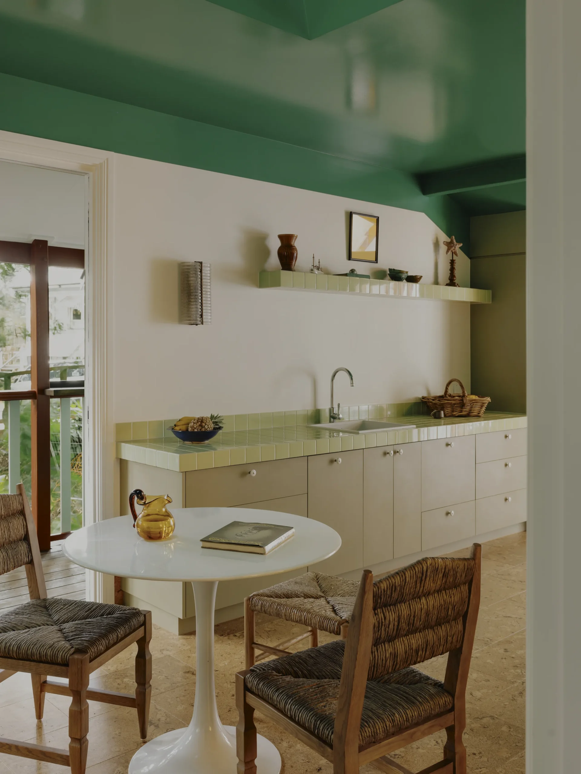 Green kitchenette with tiled counter, round table and rush-seat chairs, rainforest views through door