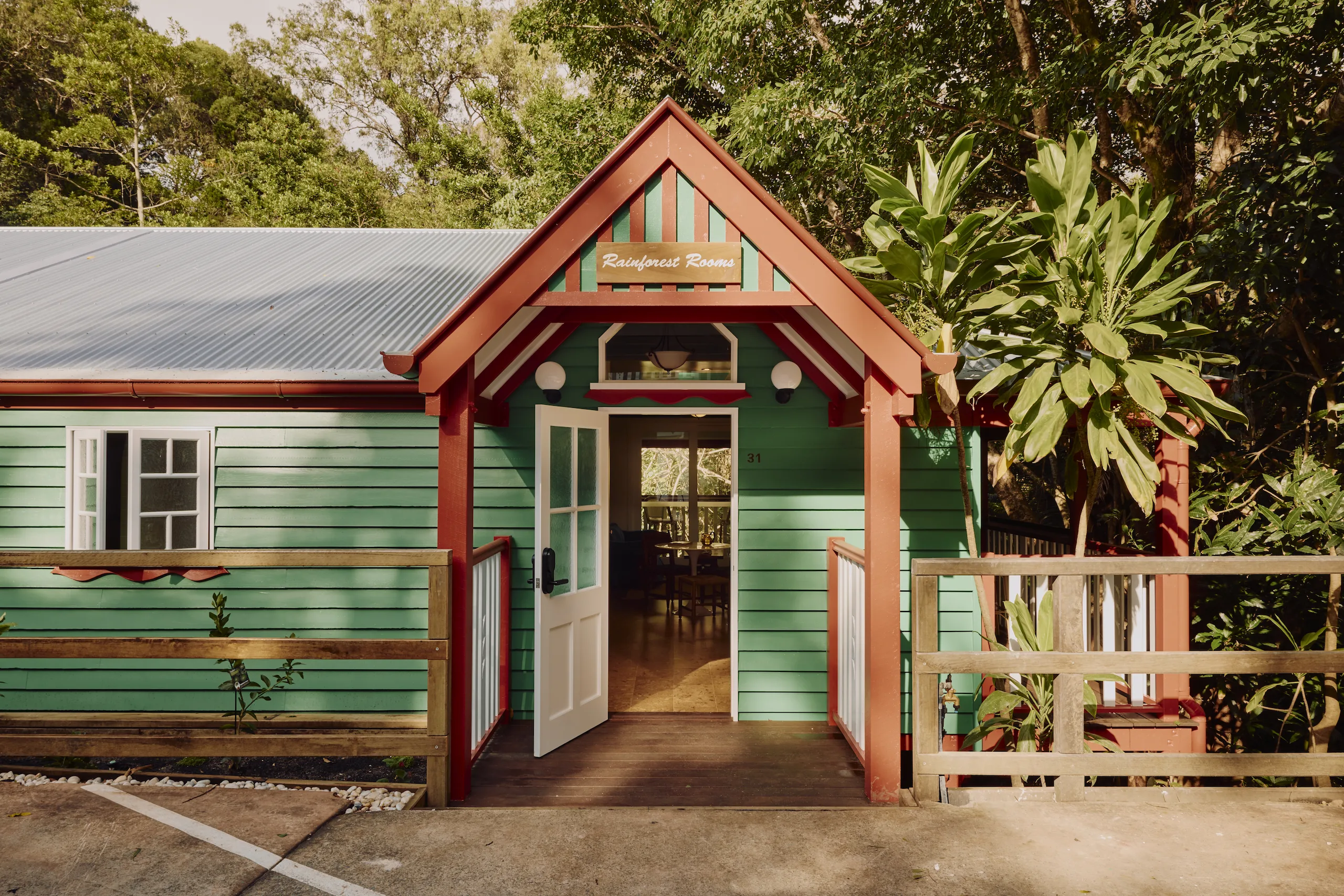Green weatherboard "Rainforest Rooms" entrance with red gabled porch, surrounded by rainforest